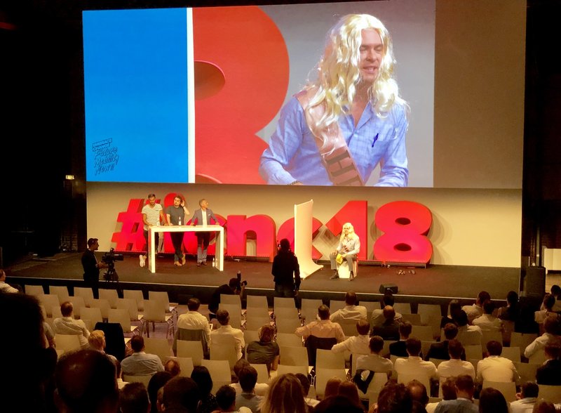 Bühne der ICNC18 mit großem roten Schriftzug Hashtag icnc18, drei Diskutanten an einem Stehtisch und einer Person mit langer blonder Perücke, auf der Leinwand dahinter im Großformat zu sehen