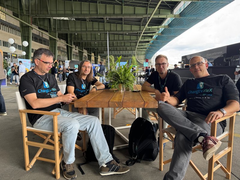 Vier Mitglieder des EV-Freaks-Teams in ChargEV-FleX-T-Shirts an einem Holztisch unter dem Hangardach des Flughafens Tempelhof